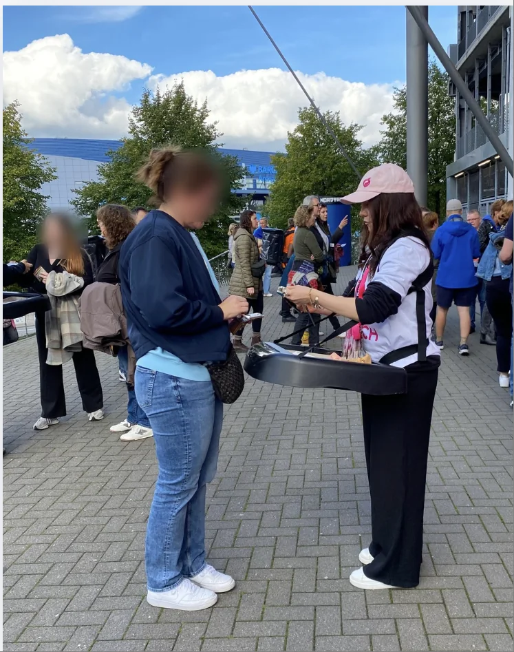 A promoter in a P3ACH shirt and cap sells edition covers from a vendor tray to a visitor outside the Volksparkstadion